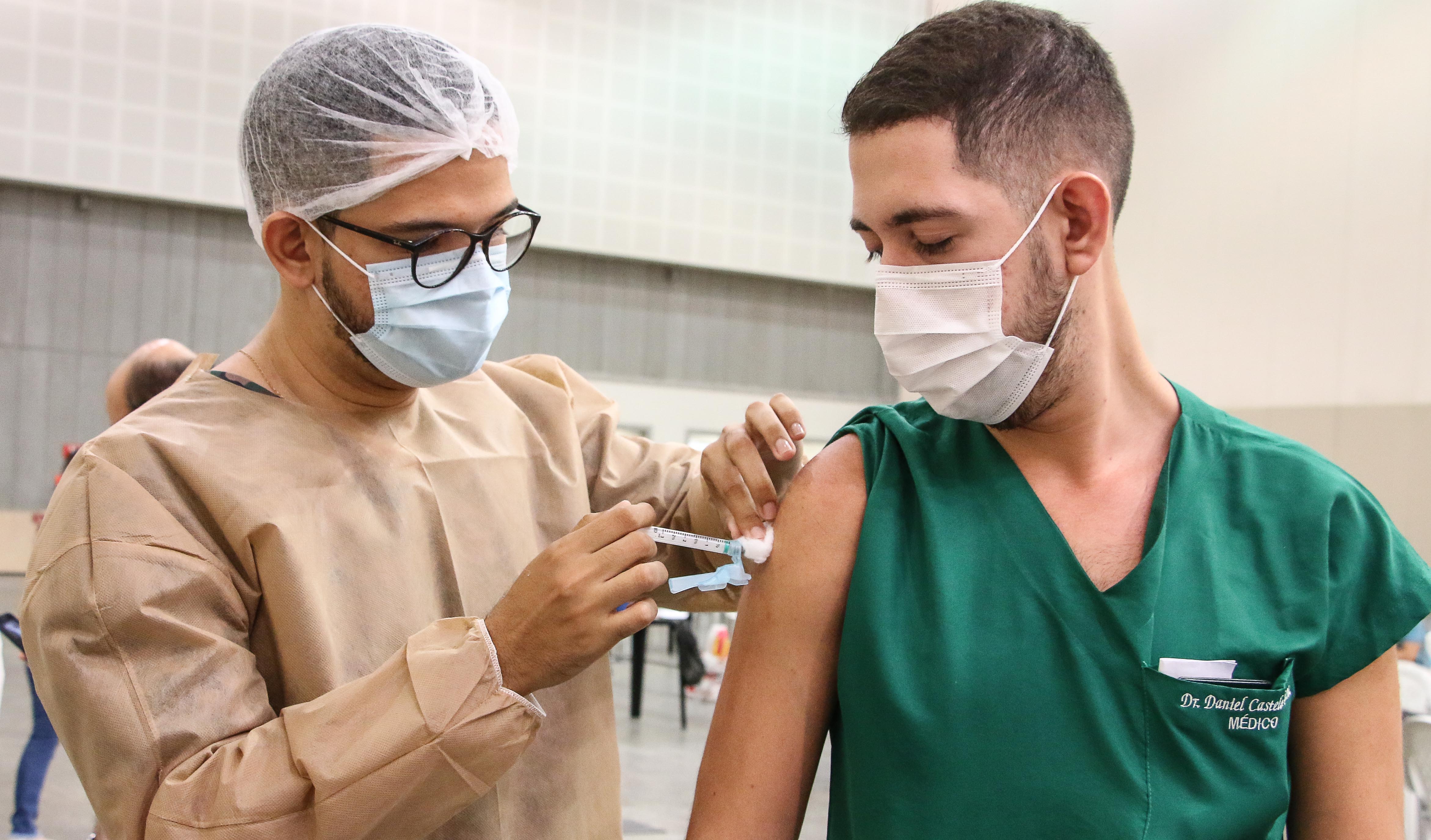 enfermeiro usando EPIs aplicando vacina em médica que usa máscara e bata com nome
